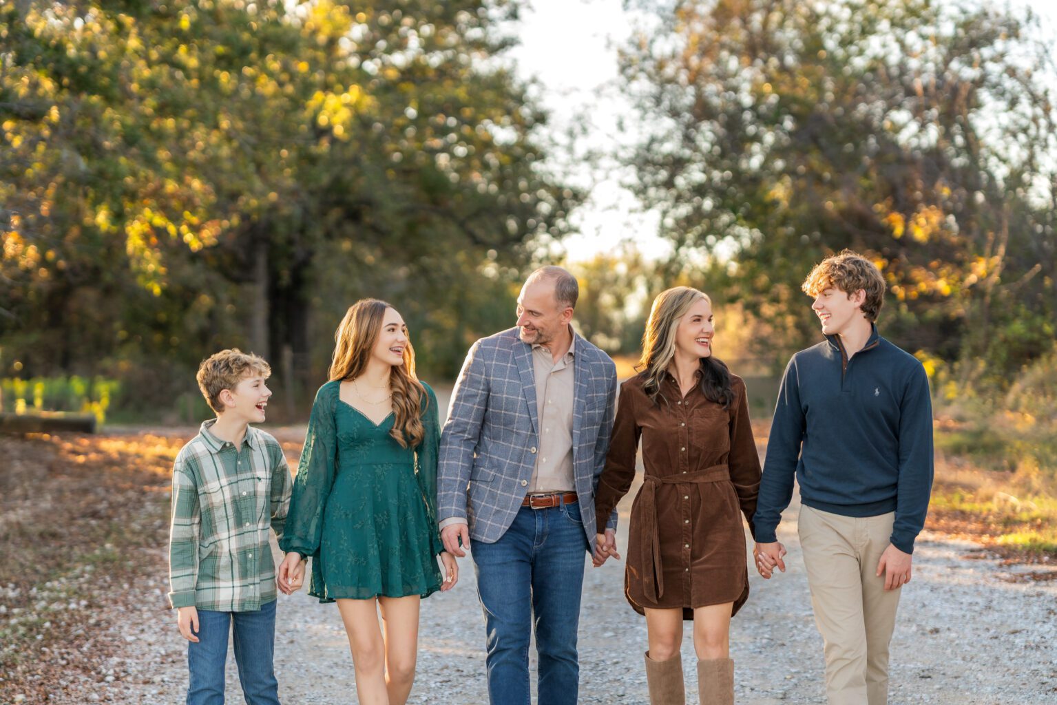 Family of five in coordinated fall outfits walking hand-in-hand and laughing together on a gravel path during a golden-hour portrait session