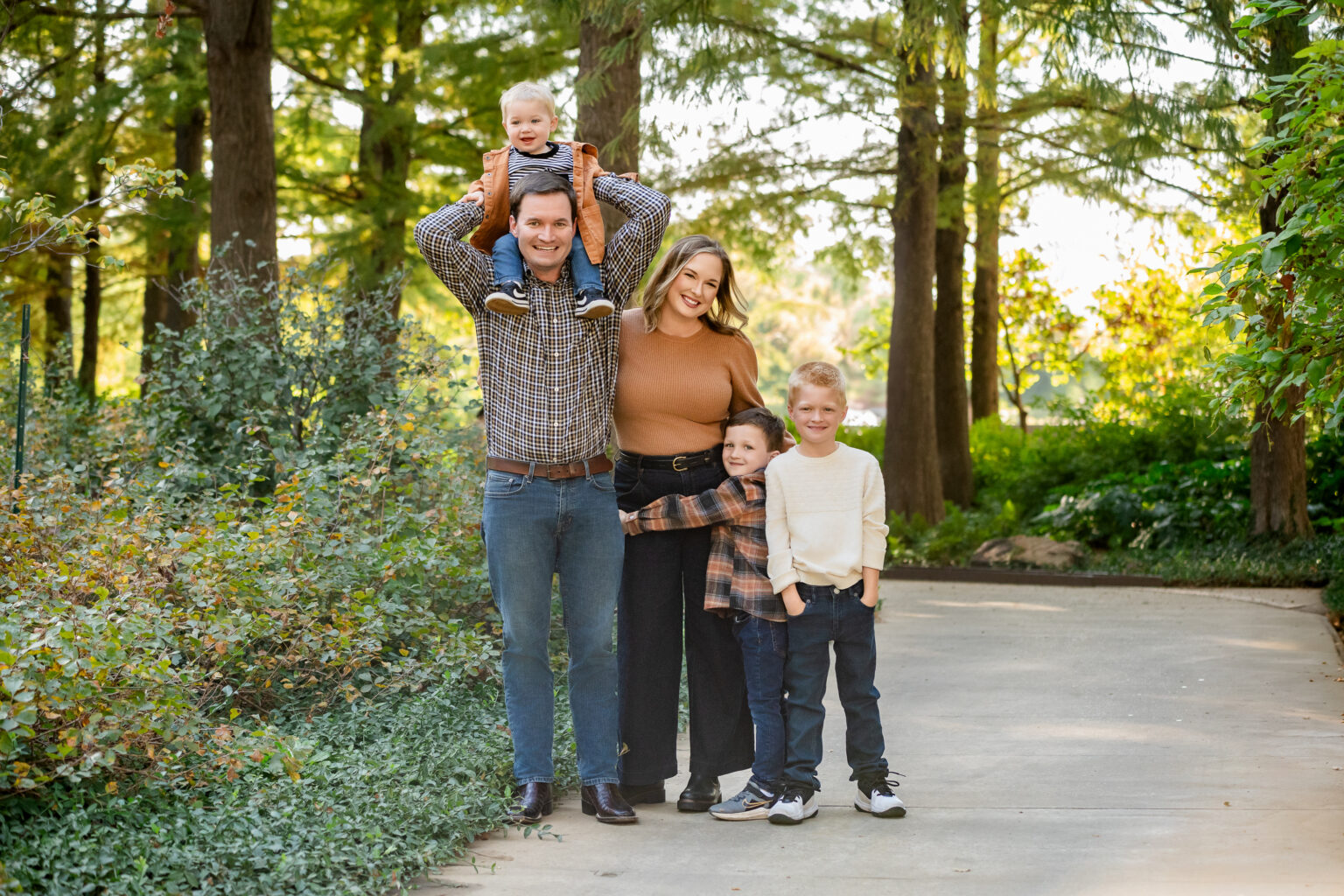 Family of five mixing plaid and solid patterns in coordinating fall colors on a wooded forest path, with a toddler riding on dad's shoulders